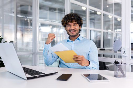 A young man, an Indian student, received good news by letter, he entered the university, passed the exam, and got a job. Sitting in the office, happy, smiling, looking at the camera.